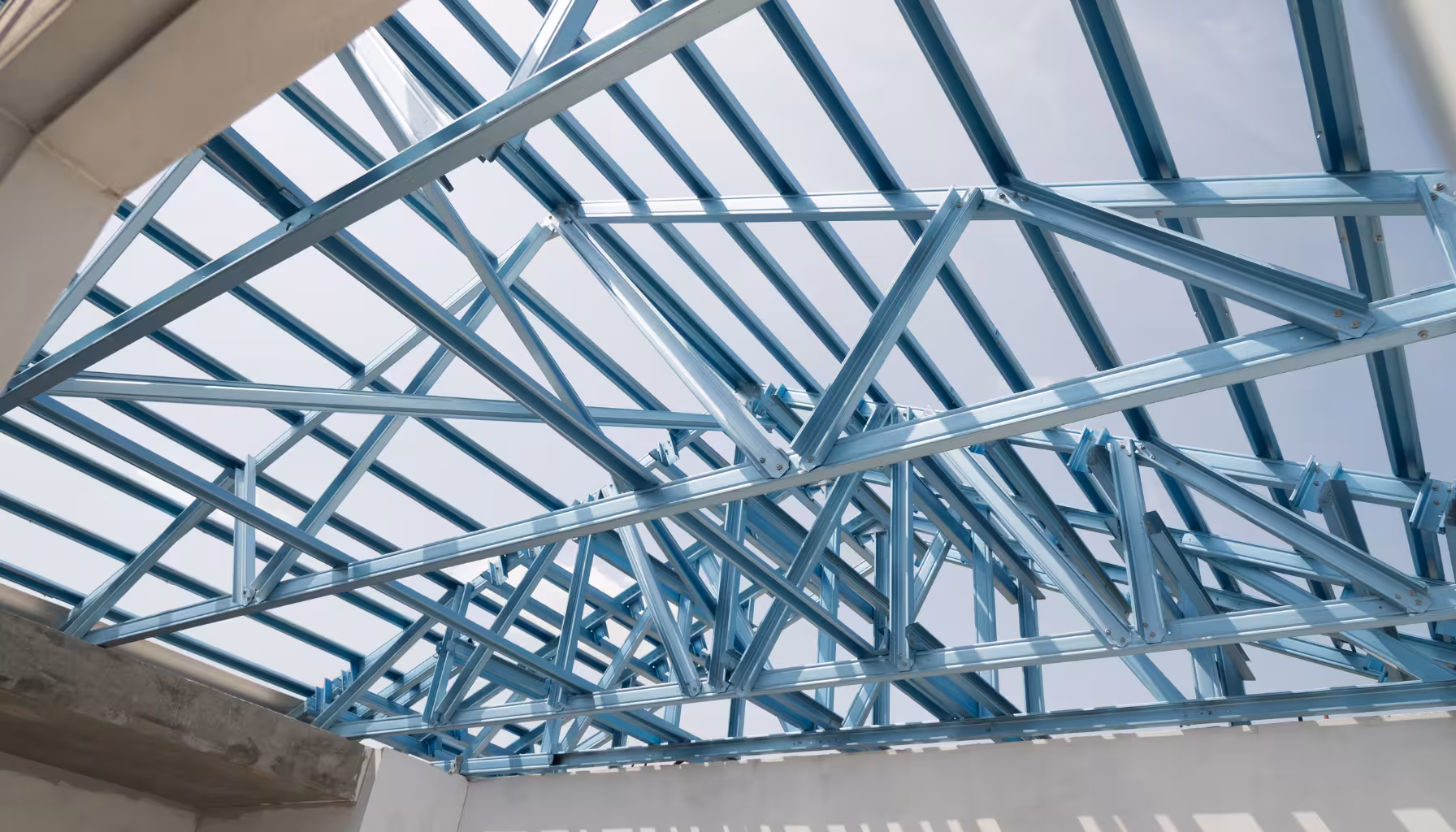 Gambar struktur rangka atap baja ringan dengan profil C yang terlihat jelas di proyek konstruksi rumah. Rangka baja ringan berwarna biru metalik terpasang rapi, menunjukkan detail truss pada atap dengan sambungan kuat dan desain modular. Langit biru di latar belakang memberi kesan terbuka dan fokus pada material baja ringan.