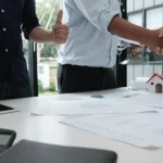Tangan berjabat dengan bangunan miniatur sebagai simbol proyek pembangunan rumah.