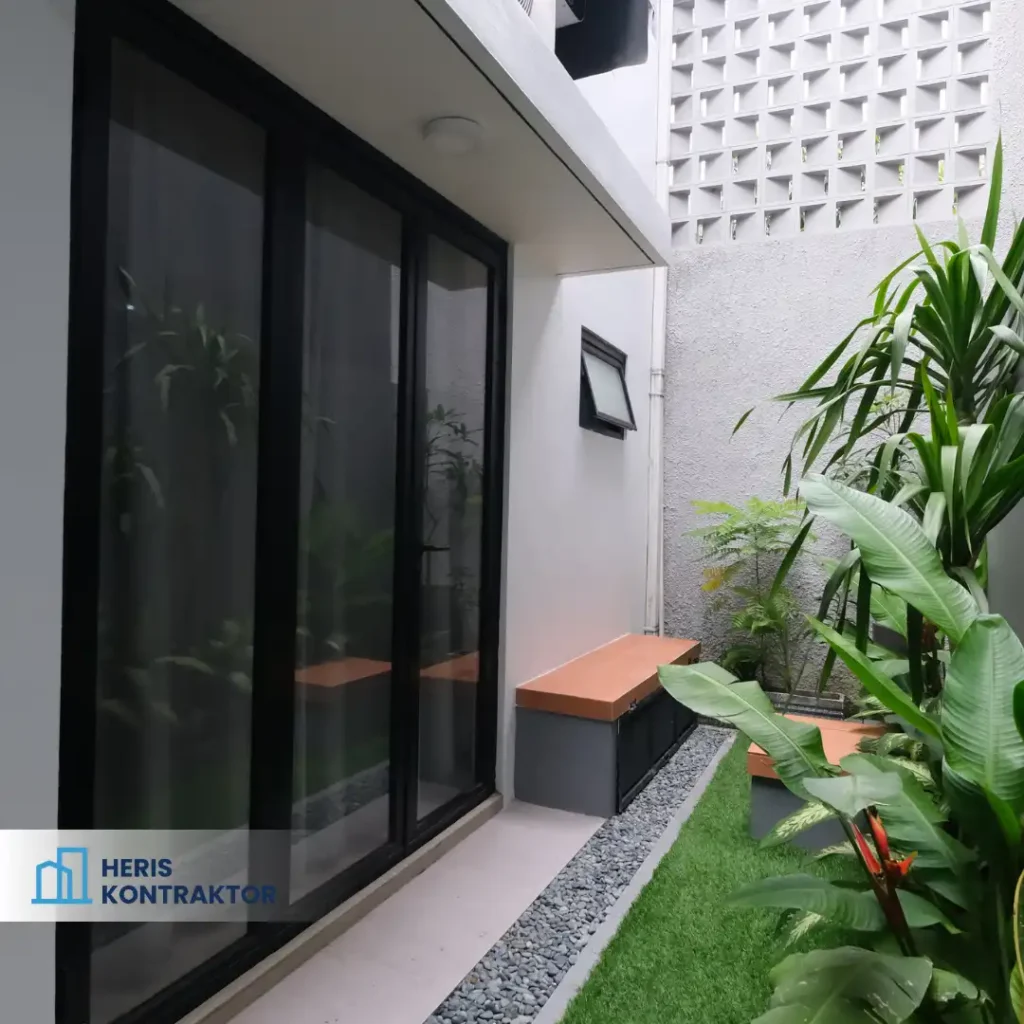Taman samping rumah dengan tanaman hijau dan bangku kayu yang nyaman.