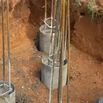Pengeboran manual pondasi strauss pile dengan lubang dan tulangan besi di lahan tanah merah.