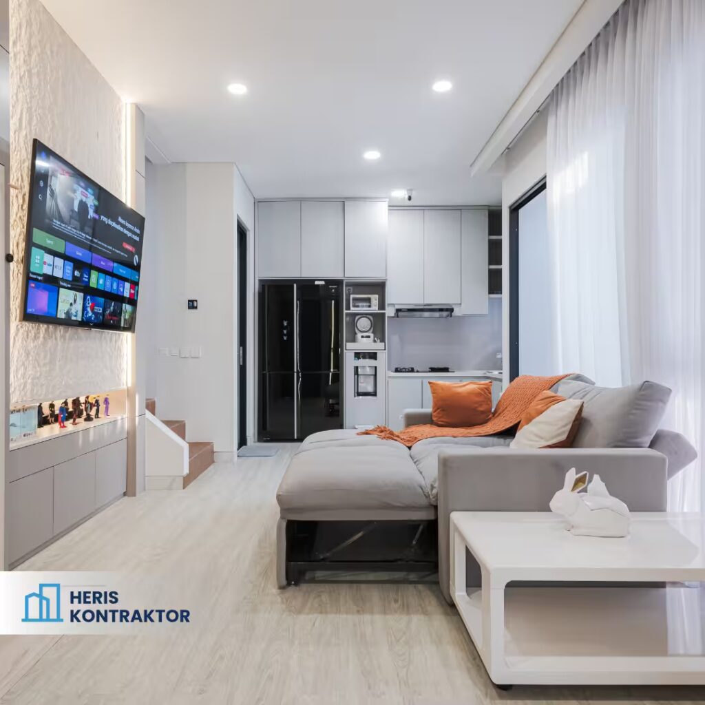 Interior ruang keluarga dengan sofa abu-abu, meja putih, backdrop TV, serta area dapur minimalis di belakang.