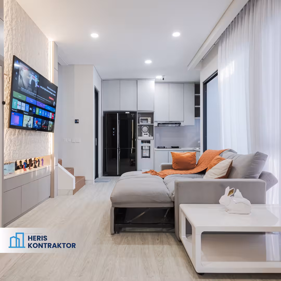 Interior ruang keluarga dengan sofa abu-abu, meja putih, backdrop TV, serta area dapur minimalis di belakang.