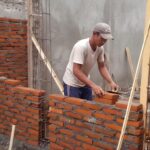 Tukang menyusun bata merah pada dinding rumah dengan pola ikatan berselang.